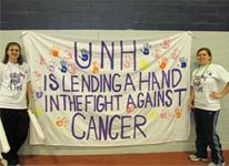 two students standing next to "UNH is lending a hand in the fight against cancer" poster