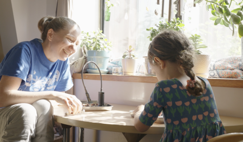 Teacher sitting at table with preschool student, smiling at child as they explore open-ended materials